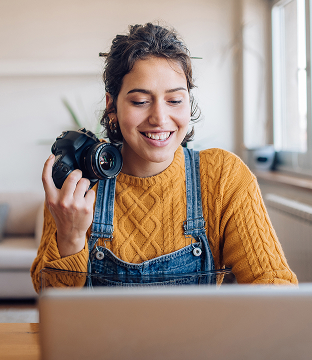 A smiling photographer holds up a camera while peering at a laptop.