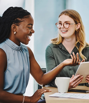 A customer and small business owner talk and smile while gathering at a table with a coffee cup, laptop and papers.