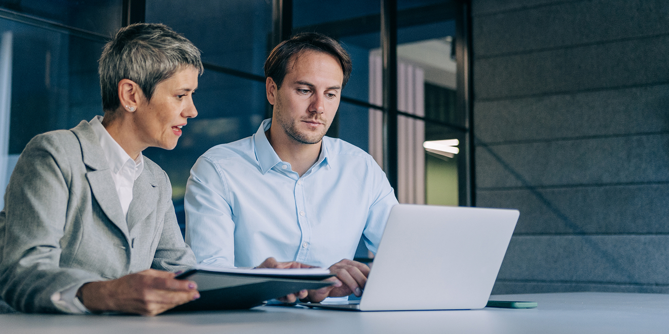 Two business people collaborate as one holds a folder and they both examine a laptop.